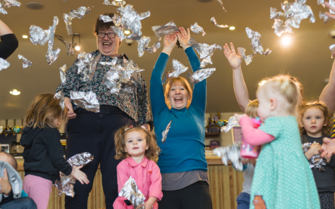 A group of adults and toddlers are standing in colourful clothes and throwing ripped up pieces of shiny silver tin foil in the air, smiling and laughing.